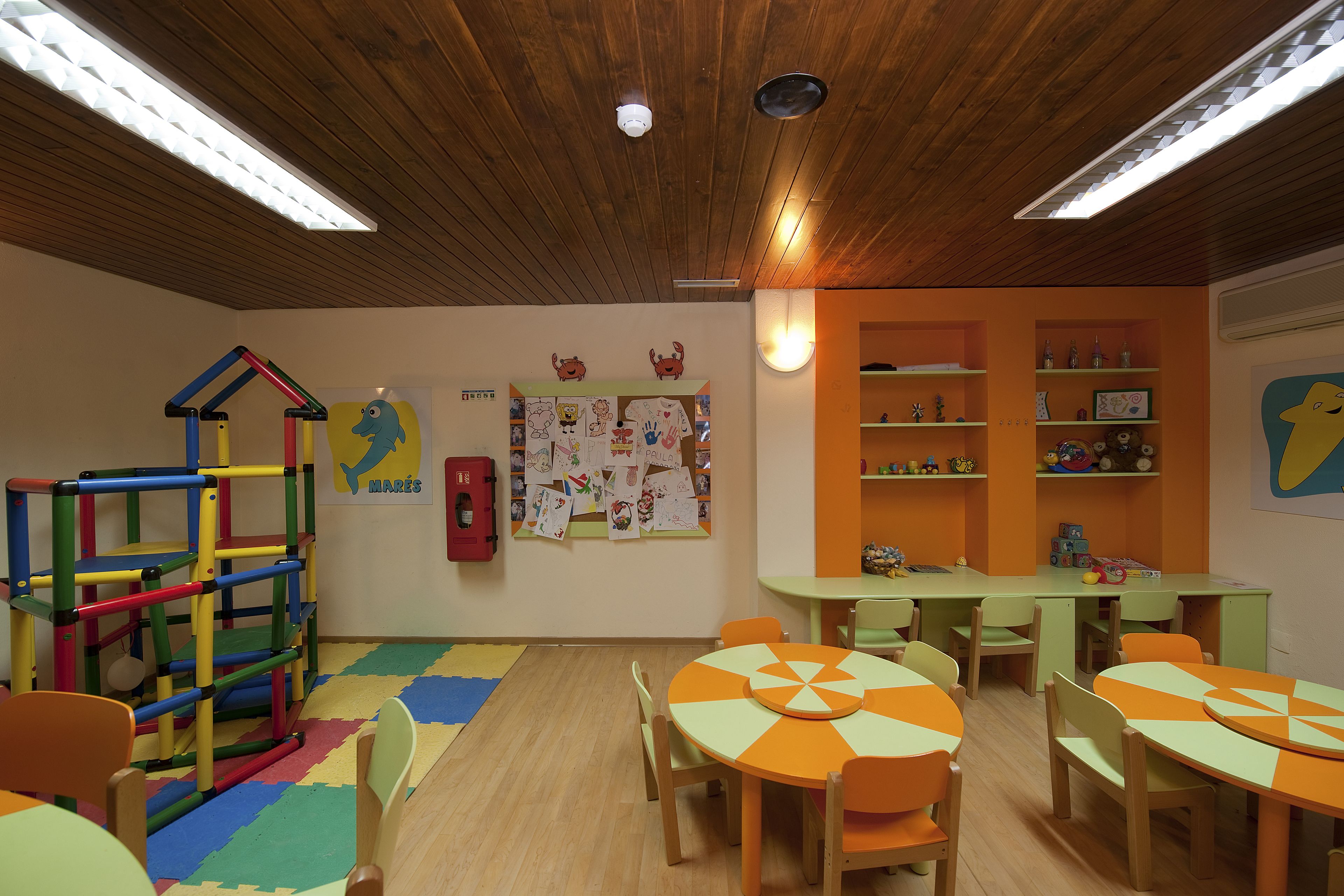 A children's playroom with colorful play structures, a bulletin board with children's drawings, and tables with orange and green tablecloths.
