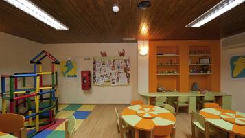 Una sala de juegos infantil con estructuras de juego coloridas, un tablón de anuncios con dibujos infantiles y mesas con manteles naranja y verde.