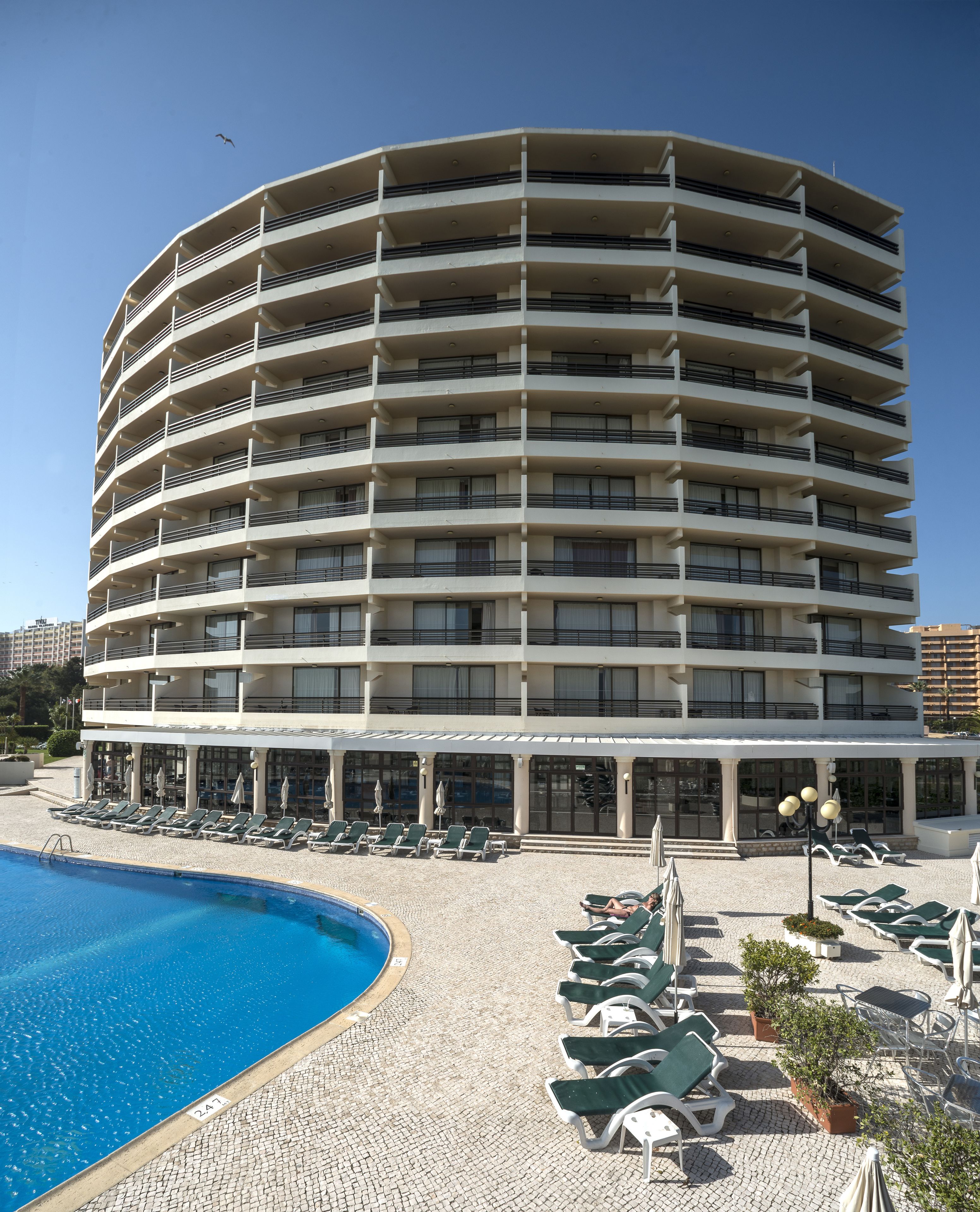 Een meerhoog hotel met een zwembadgedeelte, ligbedden en een helderblauwe lucht.