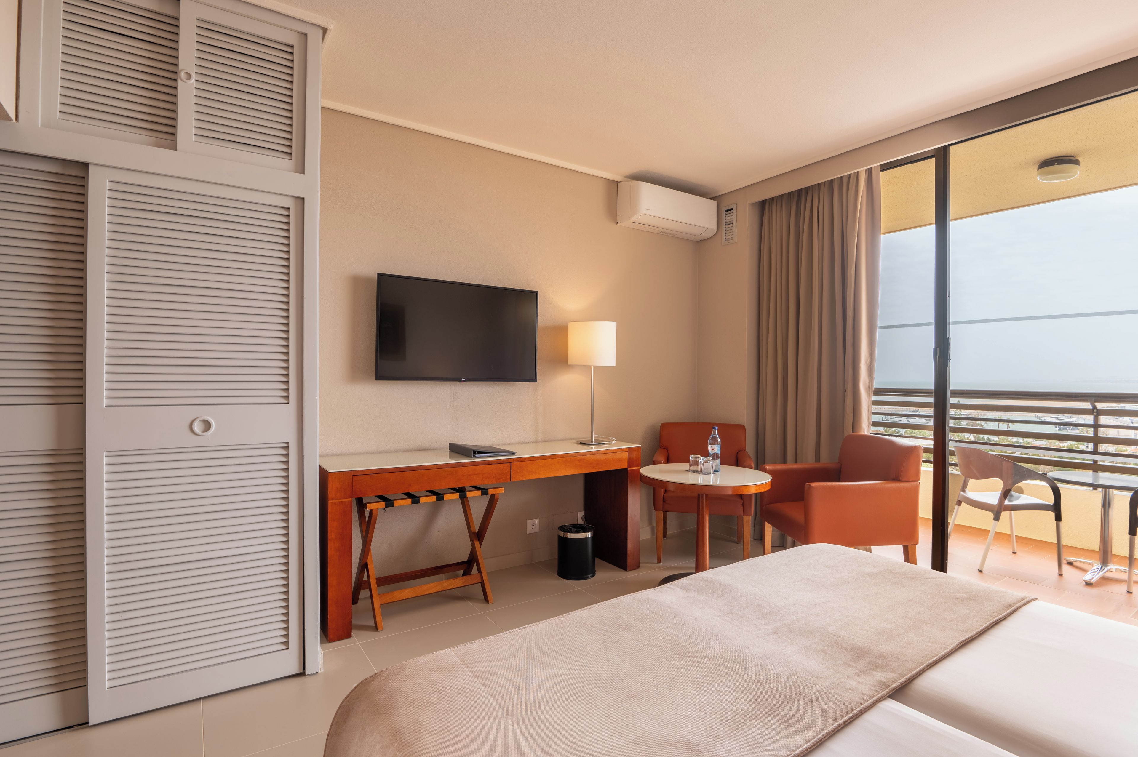 Quarto de hotel com cama, escrivaninha com cadeira, televisão, mesa pequena com cadeira e varanda com vista.