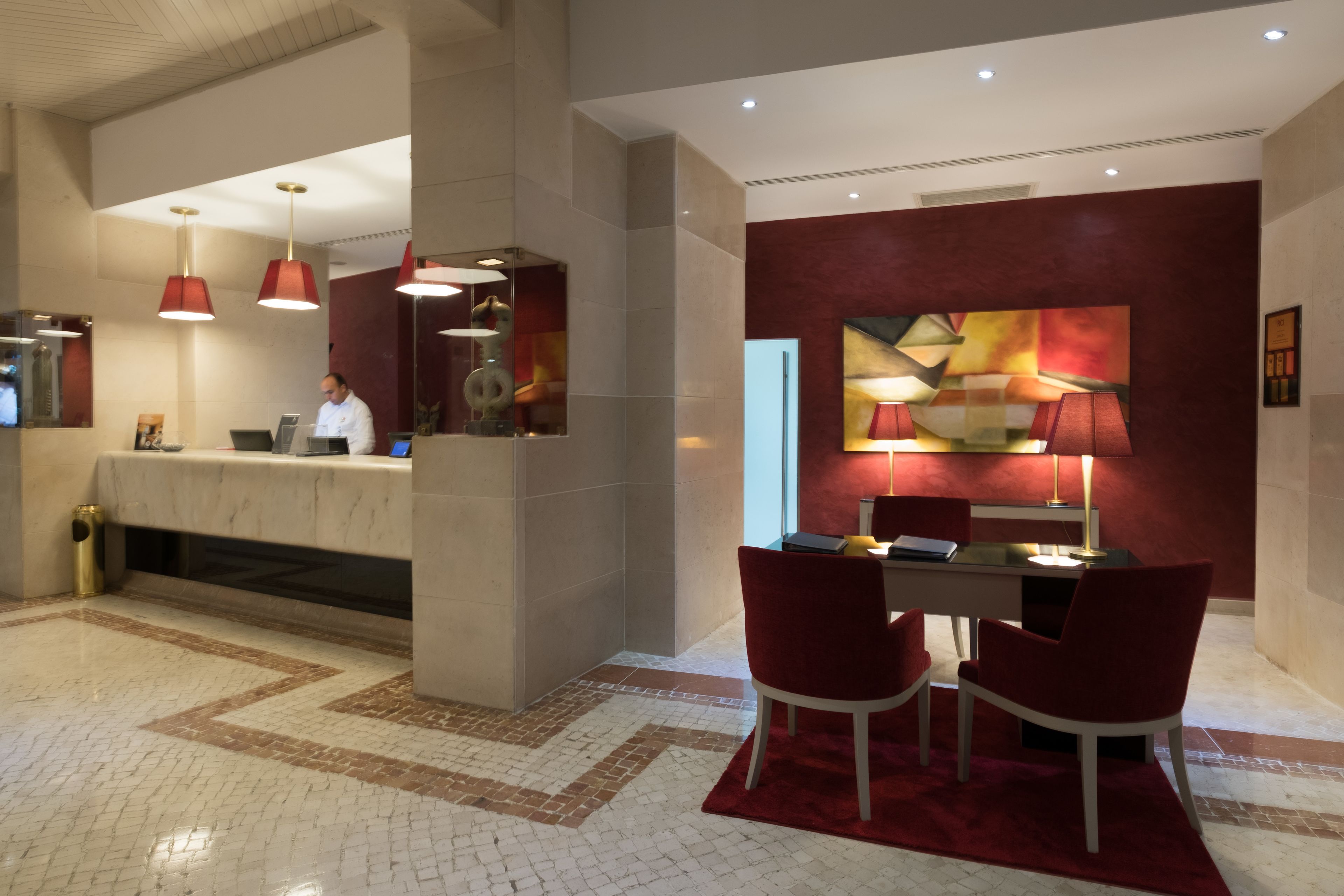 A hotel lobby with a reception desk, red chairs, a red rug, and a modern abstract painting on the wall.