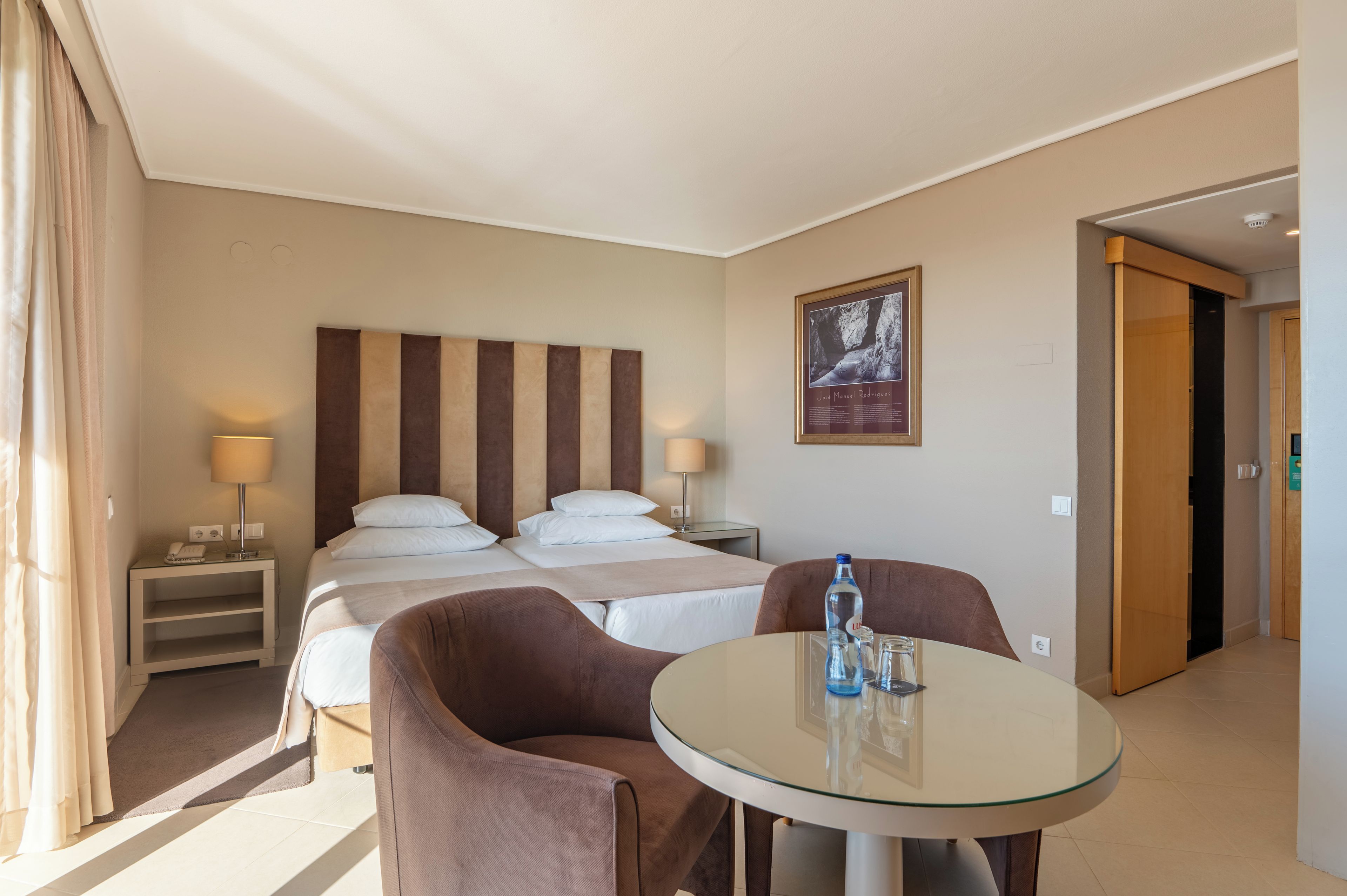 Une chambre d’hôtel avec un lit, des tables de chevet, une table en verre avec des chaises, un tableau au mur et une porte donnant sur une autre pièce.