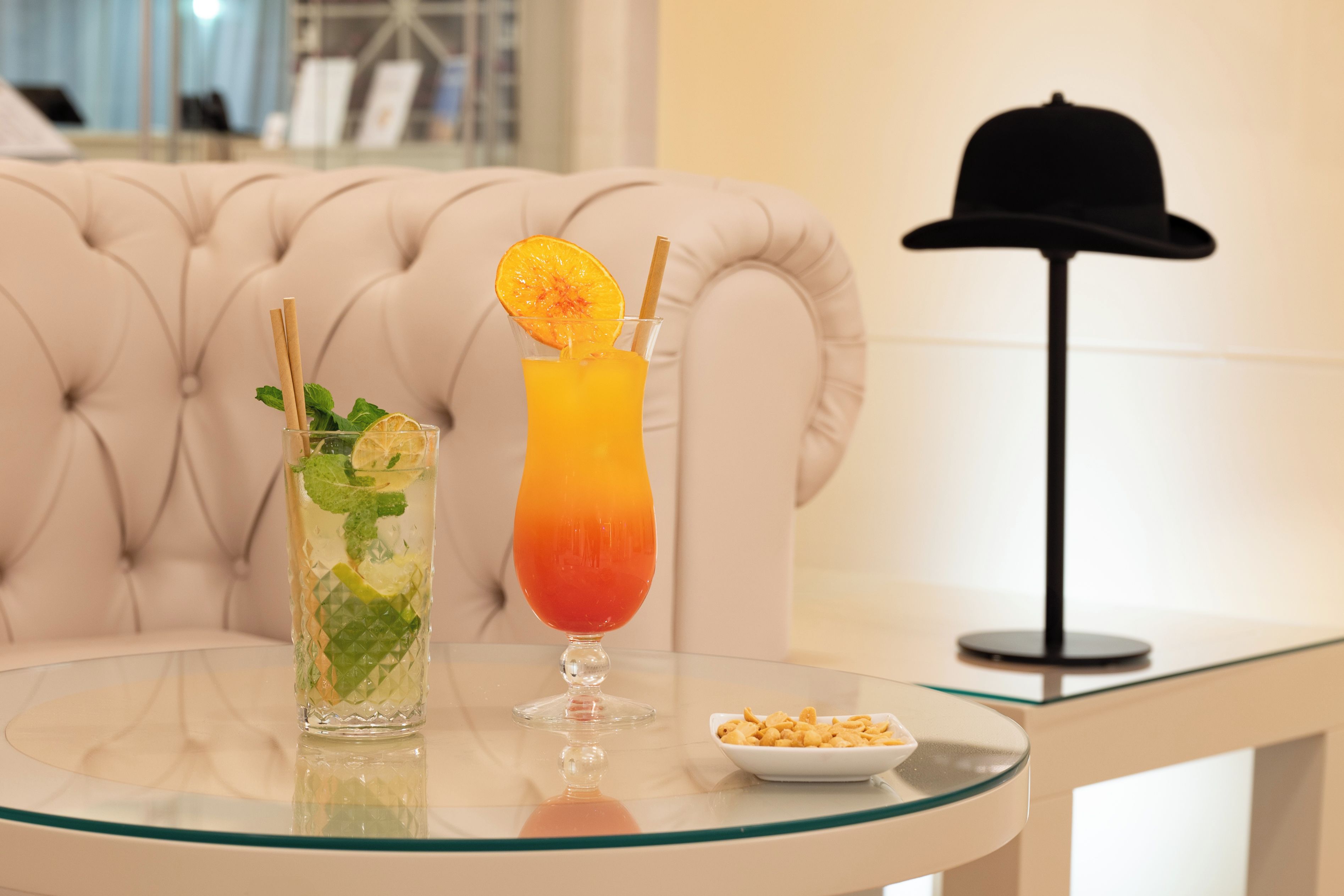 A glass table with two drinks, a chair, and a hat on a stand.