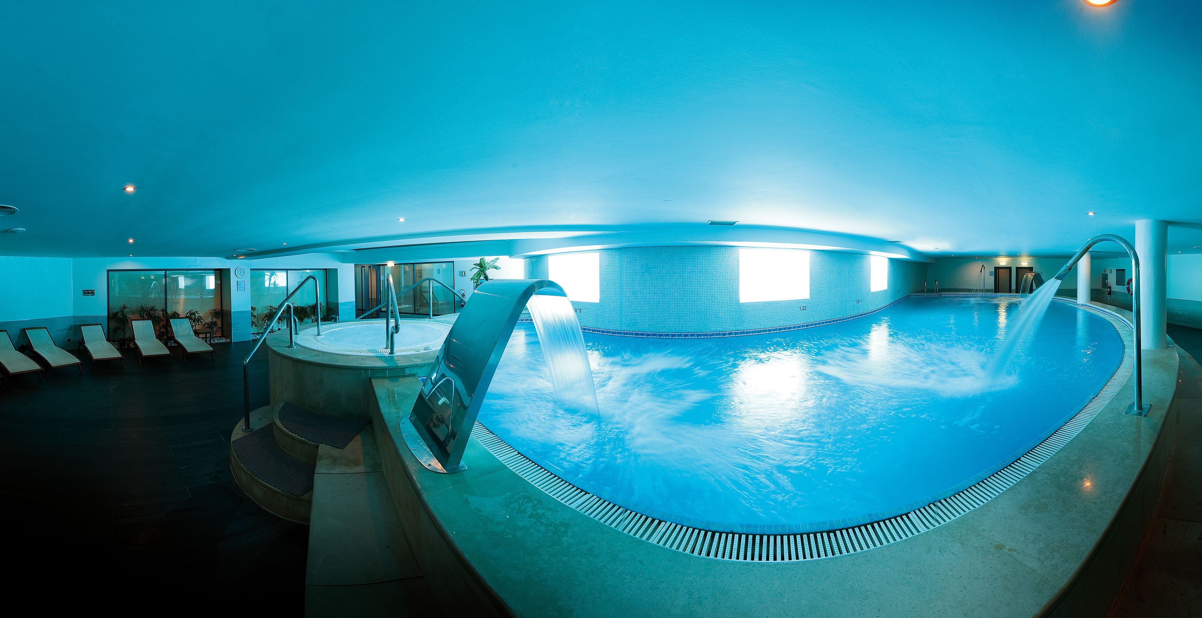 An indoor swimming pool with a curved edge, a water feature, and a sunlit area with lounge chairs.