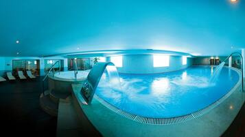 An indoor swimming pool with a curved edge, a water feature, and a sunlit area with lounge chairs.