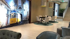 A modern hotel lobby with a mural of a tram, a dining area with round tables and chairs, and a staircase leading to an upper floor. - VIP Executive Zurique Hotel (Lisbon)