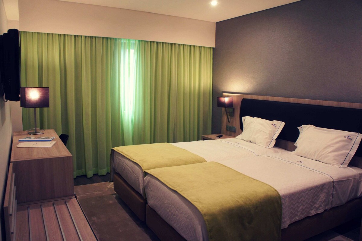 Una habitación de hotel con una cama, dos mesitas de noche, un escritorio, una lámpara y una cortina verde.