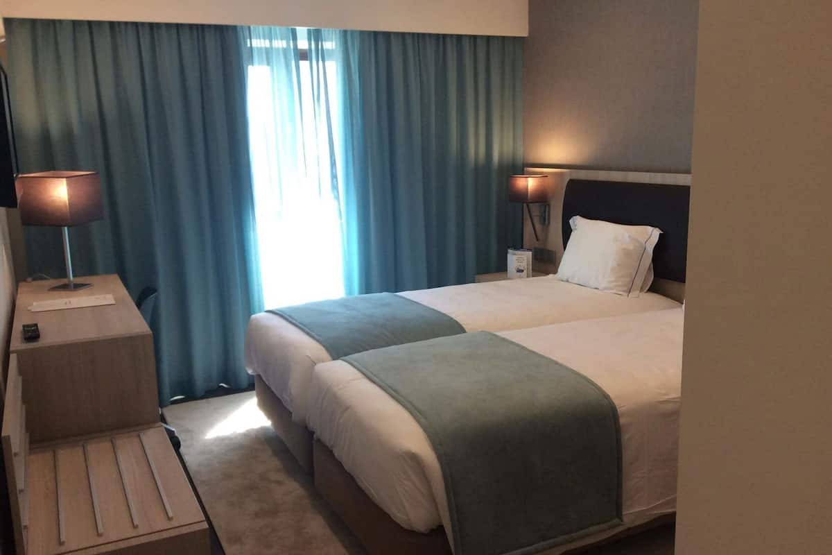 A hotel room with a large bed, two bedside tables, a lamp on each table, and a window with curtains.