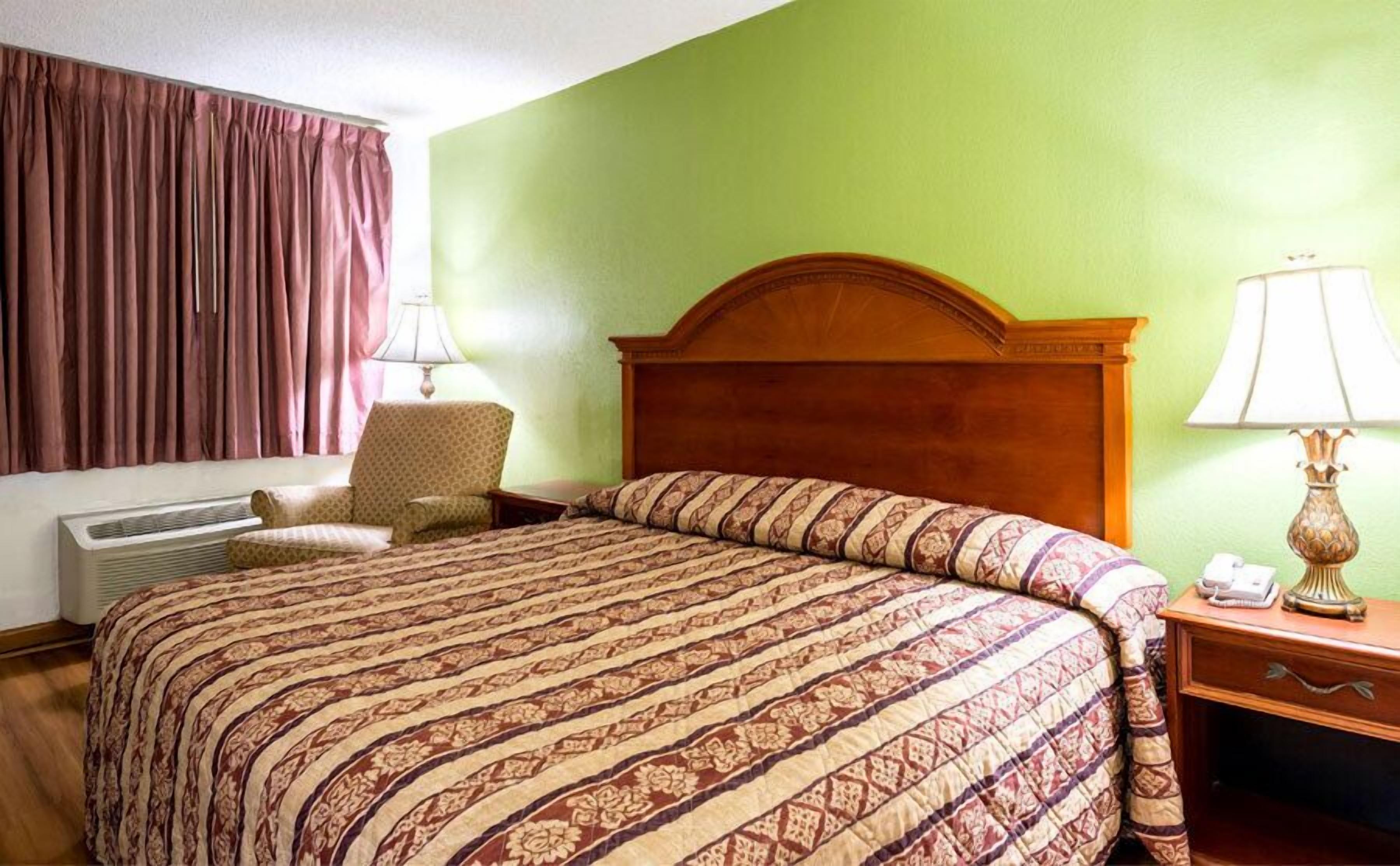 Een hotelkamer met een bed, een stoel, een nachtkastje, een lamp en een raam met gordijnen.