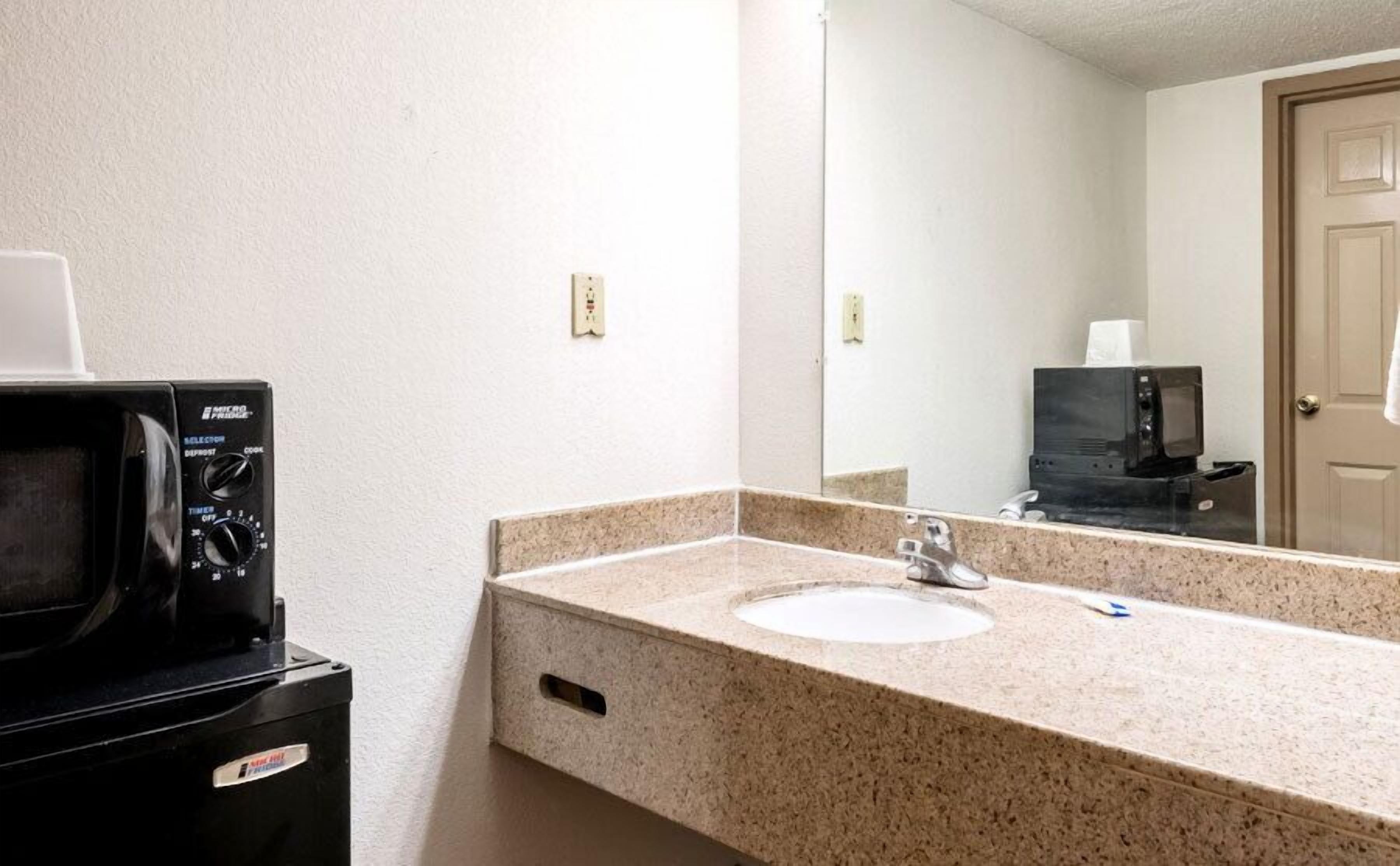 A hotel bathroom with a granite countertop sink, a mirror, a microwave, and a door.