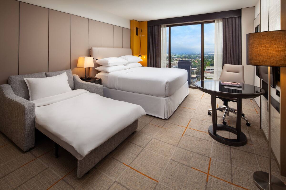 A hotel room with a large bed, a smaller bed, a sofa, a desk, and a view of the city.