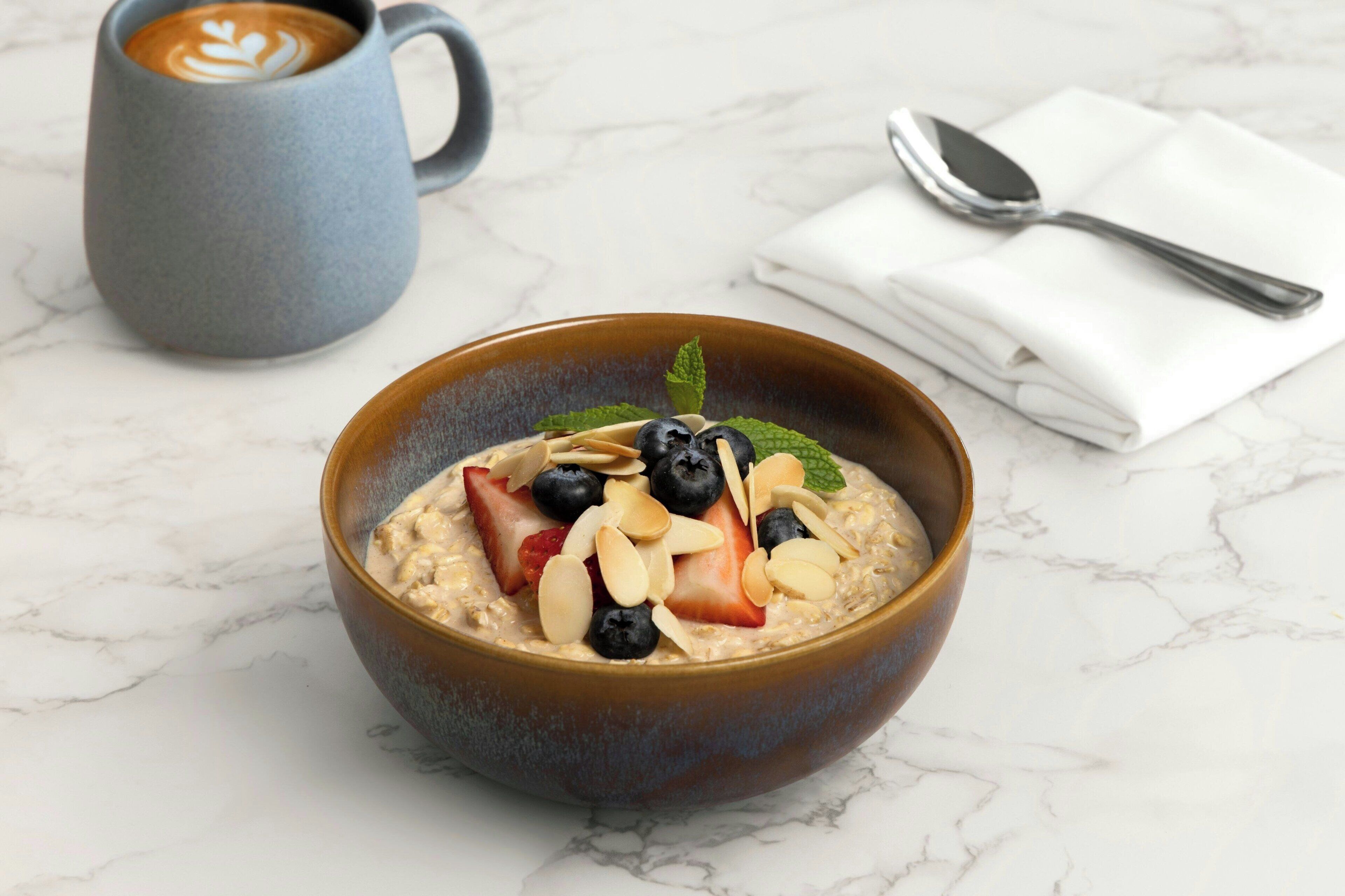 Uma tigela de aveia com frutas e nozes sobre uma superfície de mármore, uma chávena de café e uma colher sobre um guardanapo.