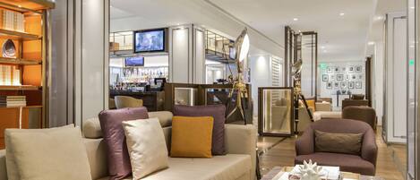 A modern hotel lobby with a sofa, armchairs, a bar area, and a television.