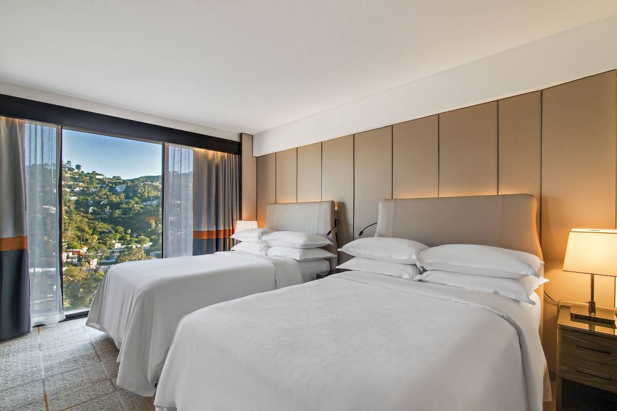 A hotel room with two beds, a large window with curtains, a nightstand with a lamp, and a view of a green landscape.