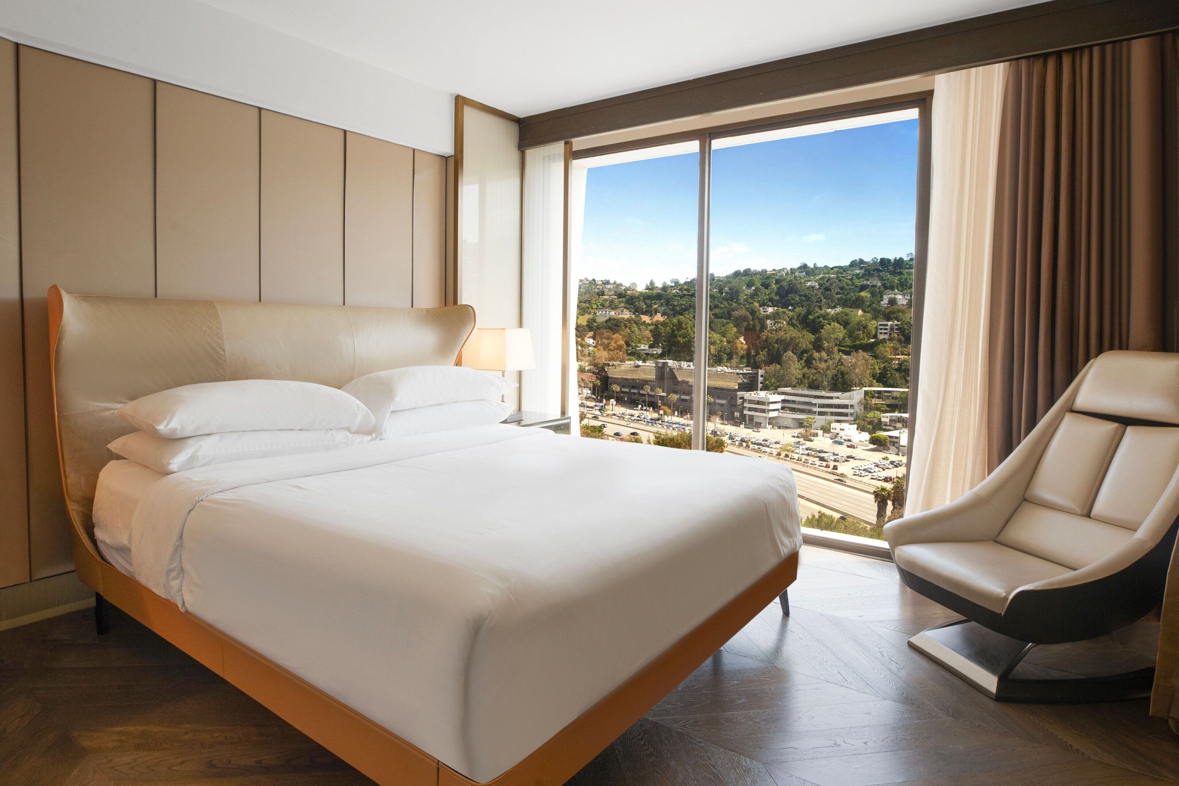 Una habitación de hotel moderna con una cama grande, una zona de estar y vistas a un paisaje urbano a través de amplios ventanales.