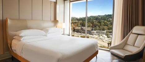 Una habitación de hotel moderna con una cama grande, una zona de estar y vistas a un paisaje urbano a través de amplios ventanales.