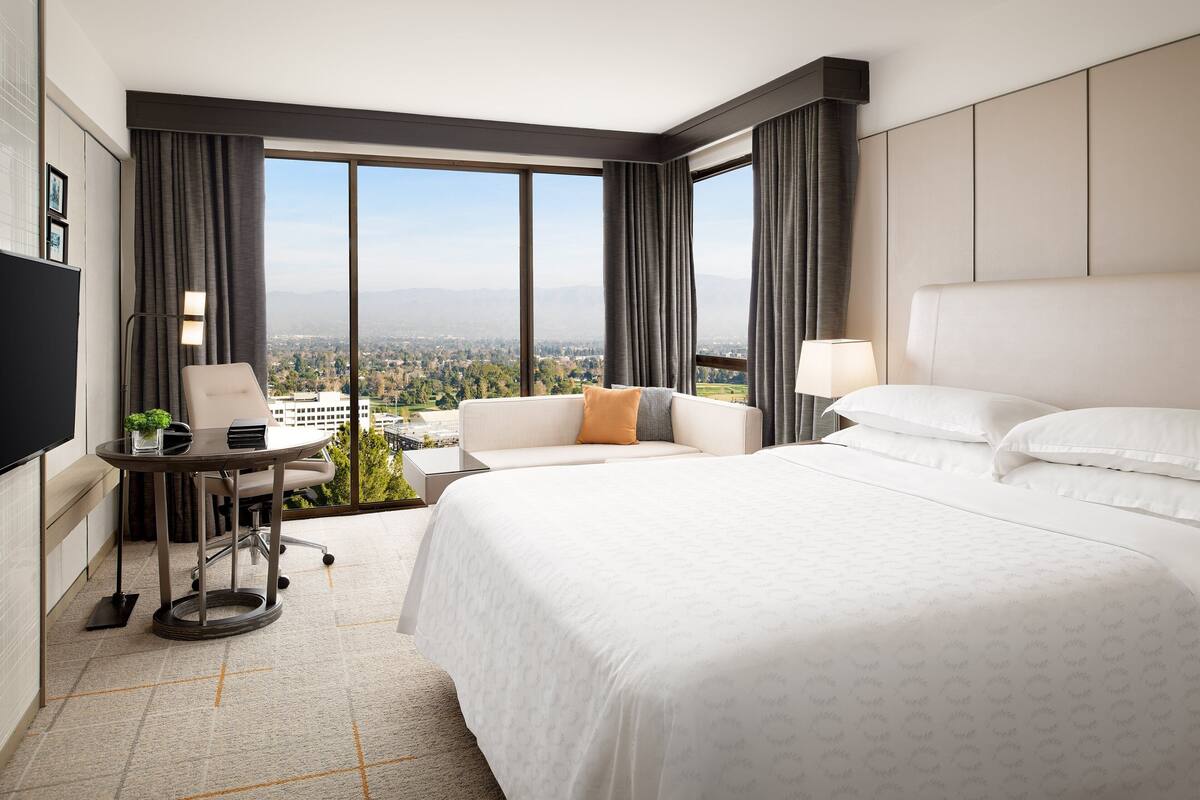 A hotel room with a large bed, a desk with a chair, a small table with a plant, and a view of the city through large windows.