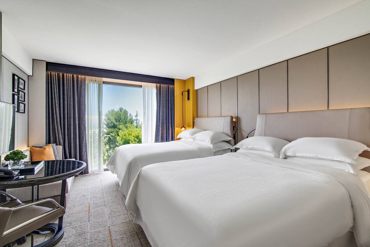 A hotel room with two beds, a desk, and a view of greenery.
