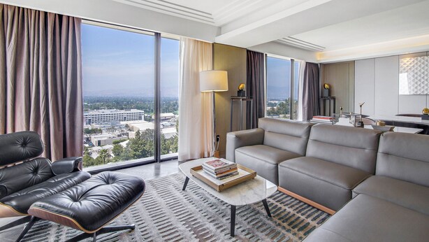 A modern hotel room with a large window, a leather sofa, a coffee table with books, and a view of the cityscape. - Sheraton Universal Hotel (Universal City)