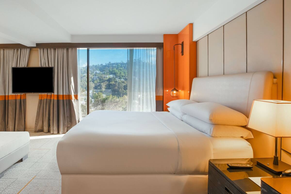 A hotel room with a large bed, a TV, a desk with a lamp, and a view of the city.