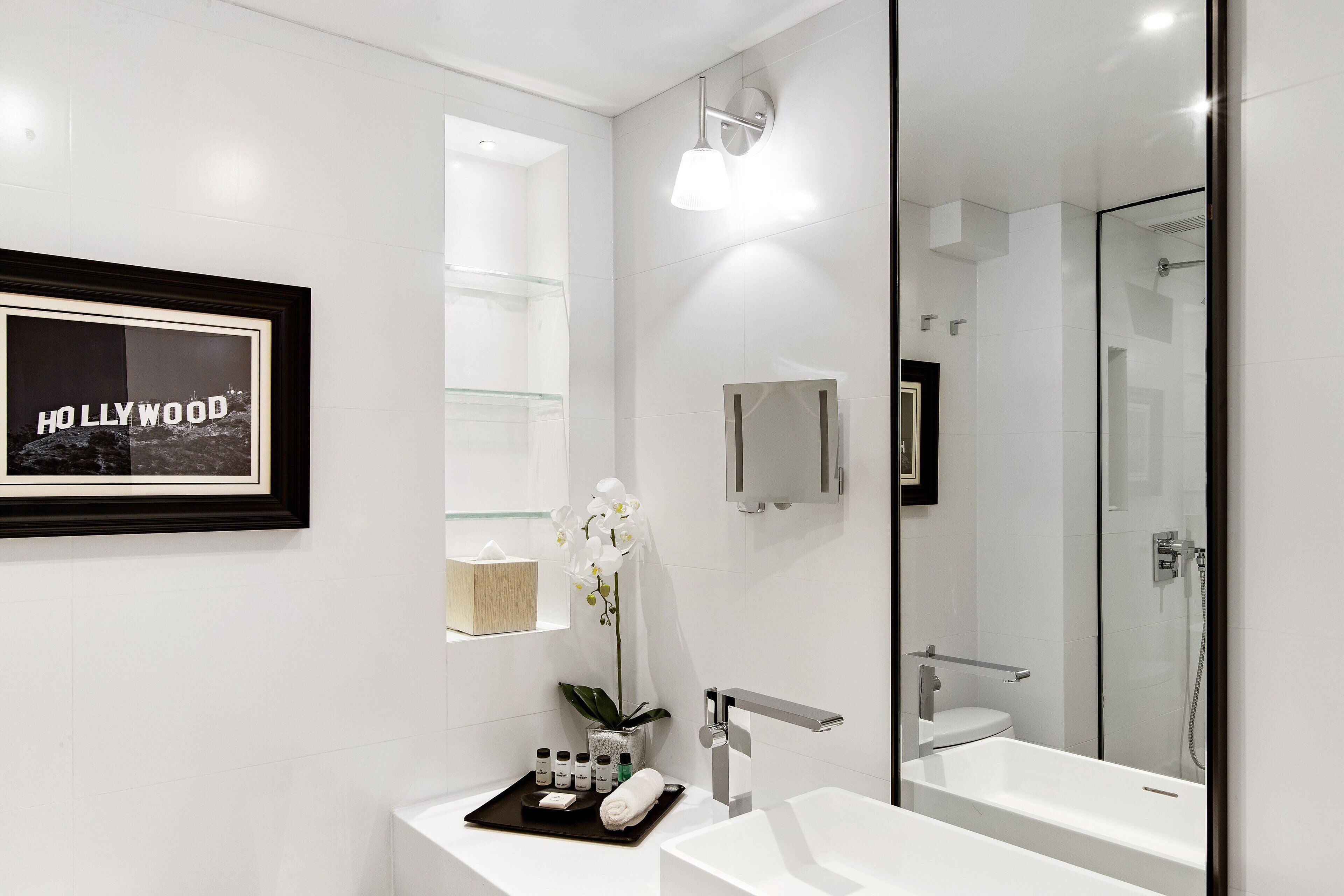 Un baño moderno con un espejo grande, un cuadro en la pared, cabina de ducha de vidrio y un lavabo blanco con artículos de aseo.