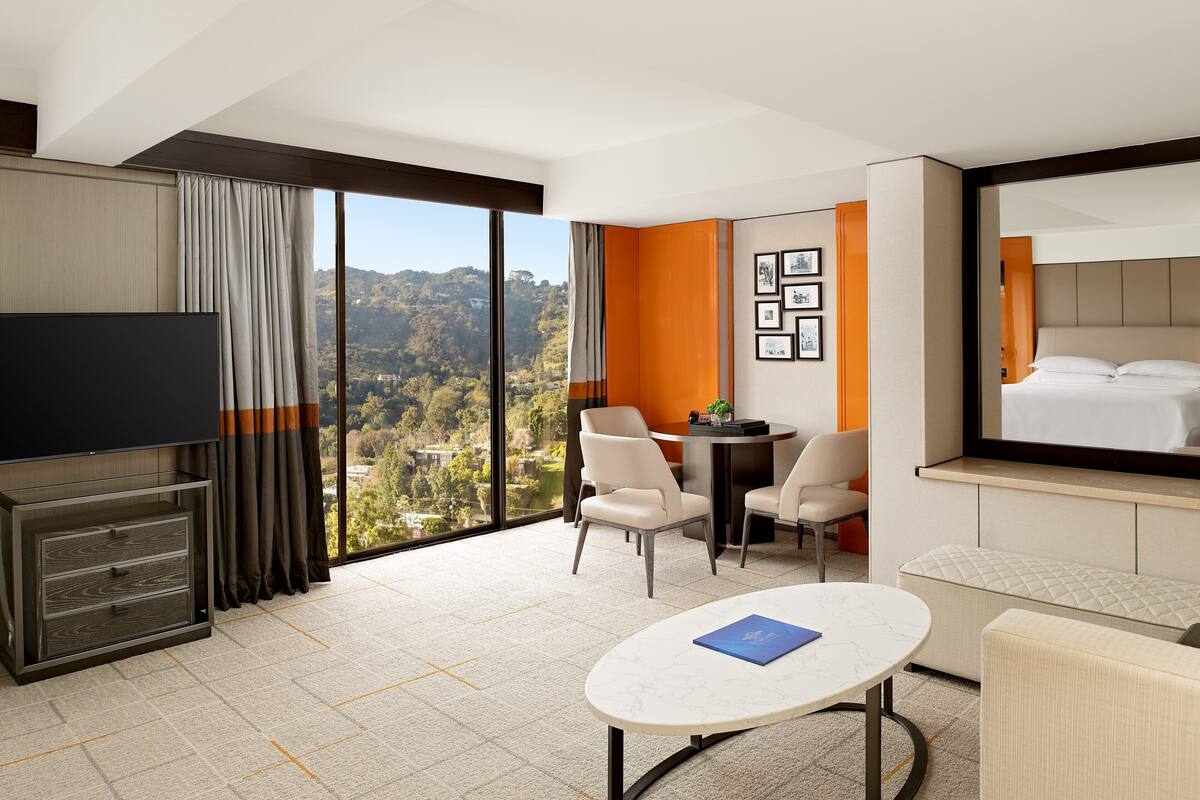 A hotel room with a large window, a flat-screen TV, a dining table, and a comfortable seating area.