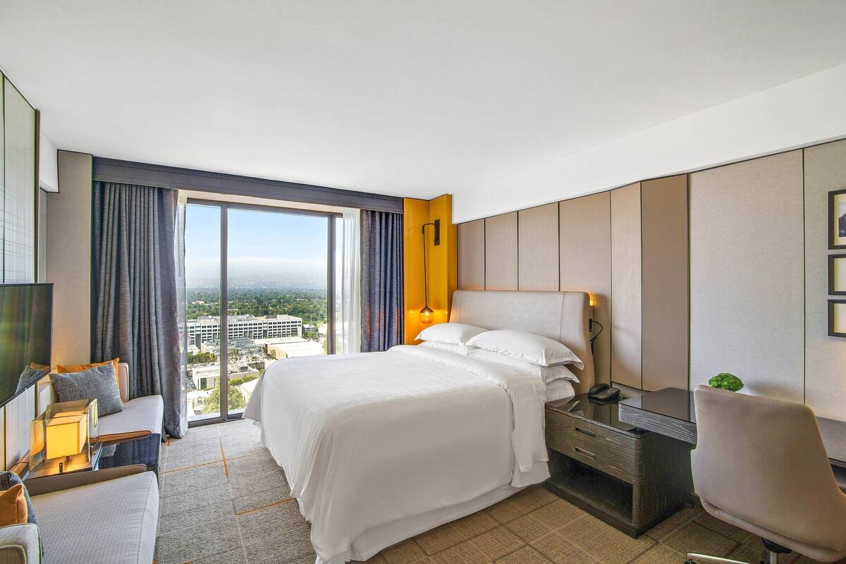 A hotel room with a large bed, a desk, a chair, a TV, and a view of the city through the window.