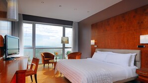 A hotel room with a large bed, wooden floor, a desk with a TV, and a view of the sea through large windows. - Hotel Re-Ndama (Libreville)