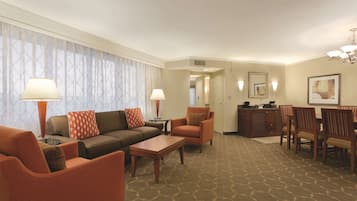 A hotel room with a living area, dining table, and chairs. There is a sofa, armchairs, and a coffee table. The room has a patterned carpet and a chandelier.