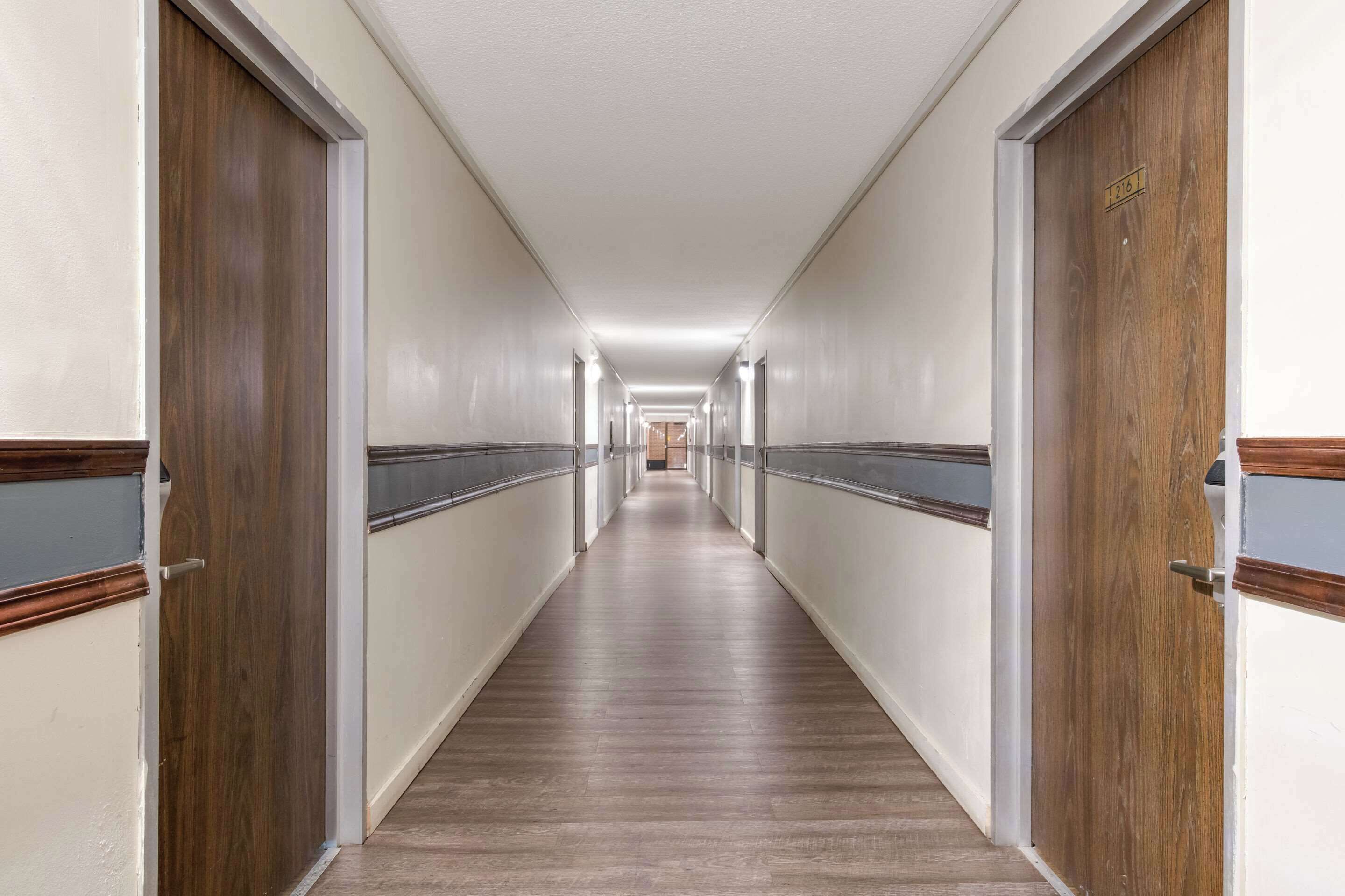 Un largo y estrecho pasillo de hotel con puertas de madera a ambos lados, suelo de color claro e iluminación empotrada en el techo.