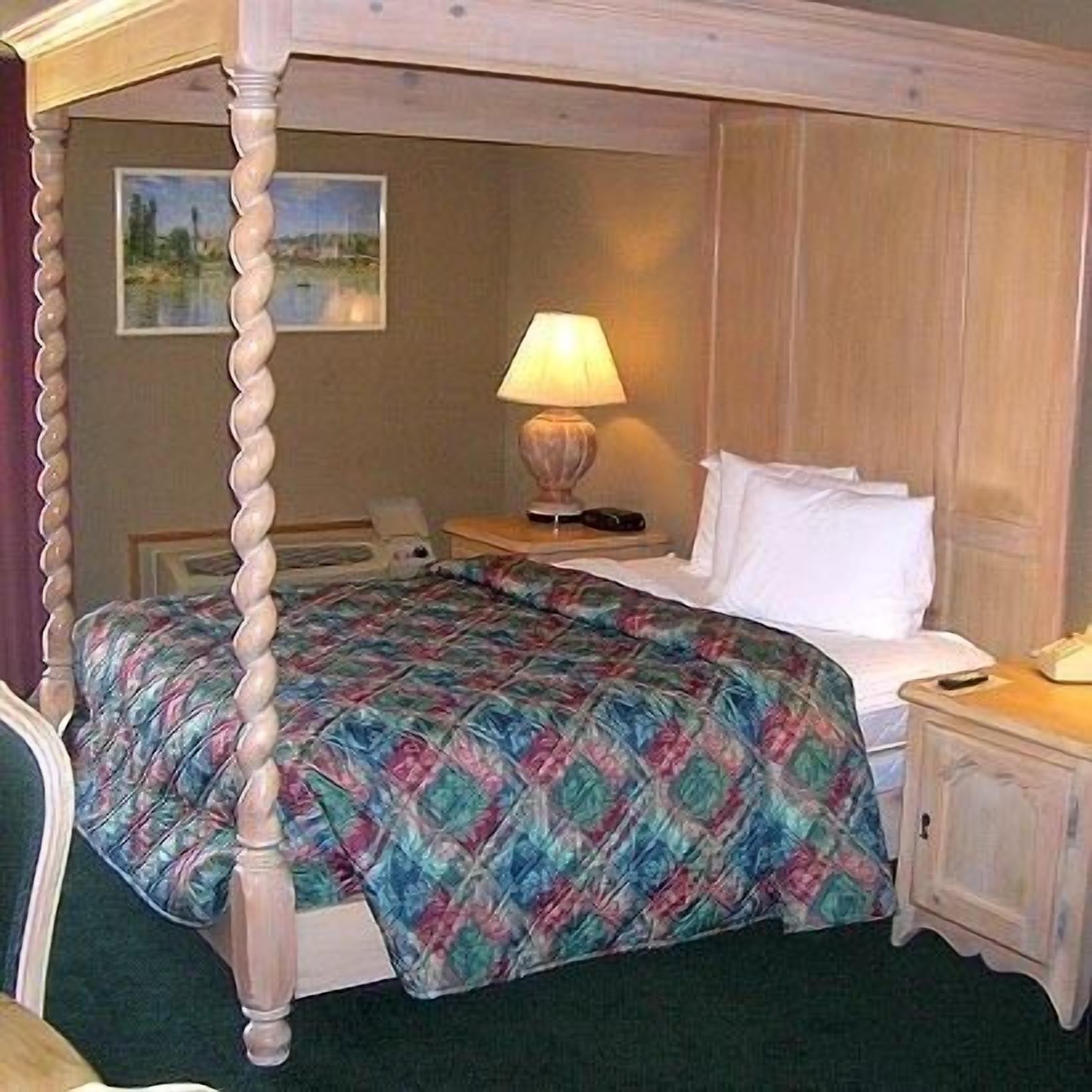 A four-poster bed with a colorful quilt, a bedside lamp, a nightstand, and a painting on the wall.