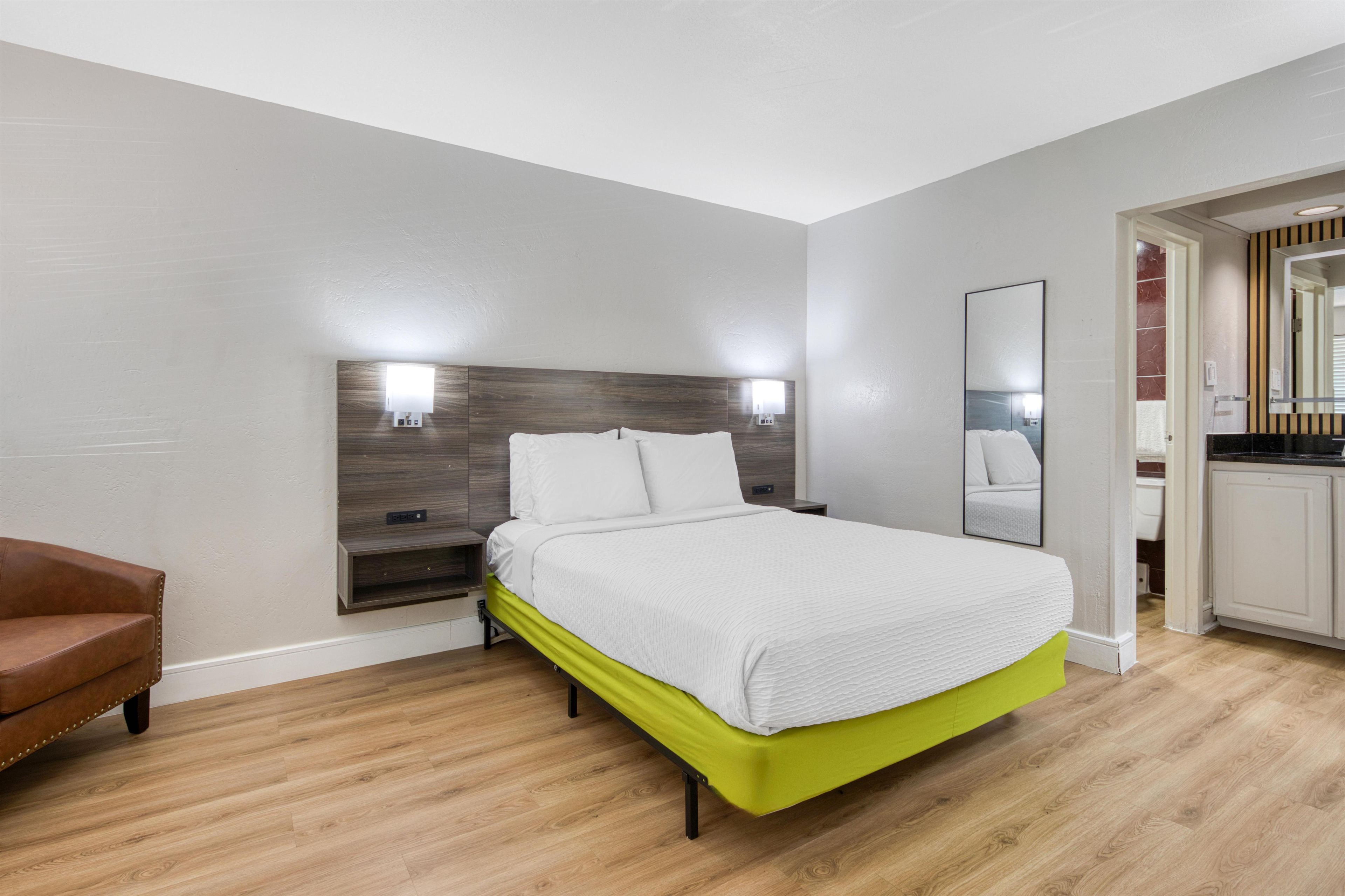 A hotel room with a bed, a bedside table, a chair, a mirror, and a kitchenette area.