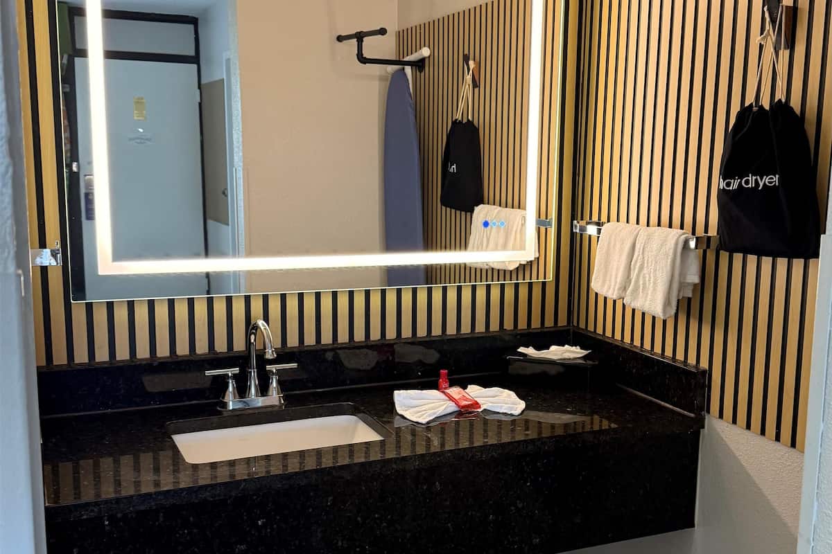 A modern bathroom with a black granite countertop, a large mirror with LED lighting, a towel rack with a white towel, and a black bag hanging on the wall.