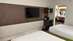 A hotel room with two beds, a TV mounted on the wall, a small kitchenette, and a mirror reflecting the room.