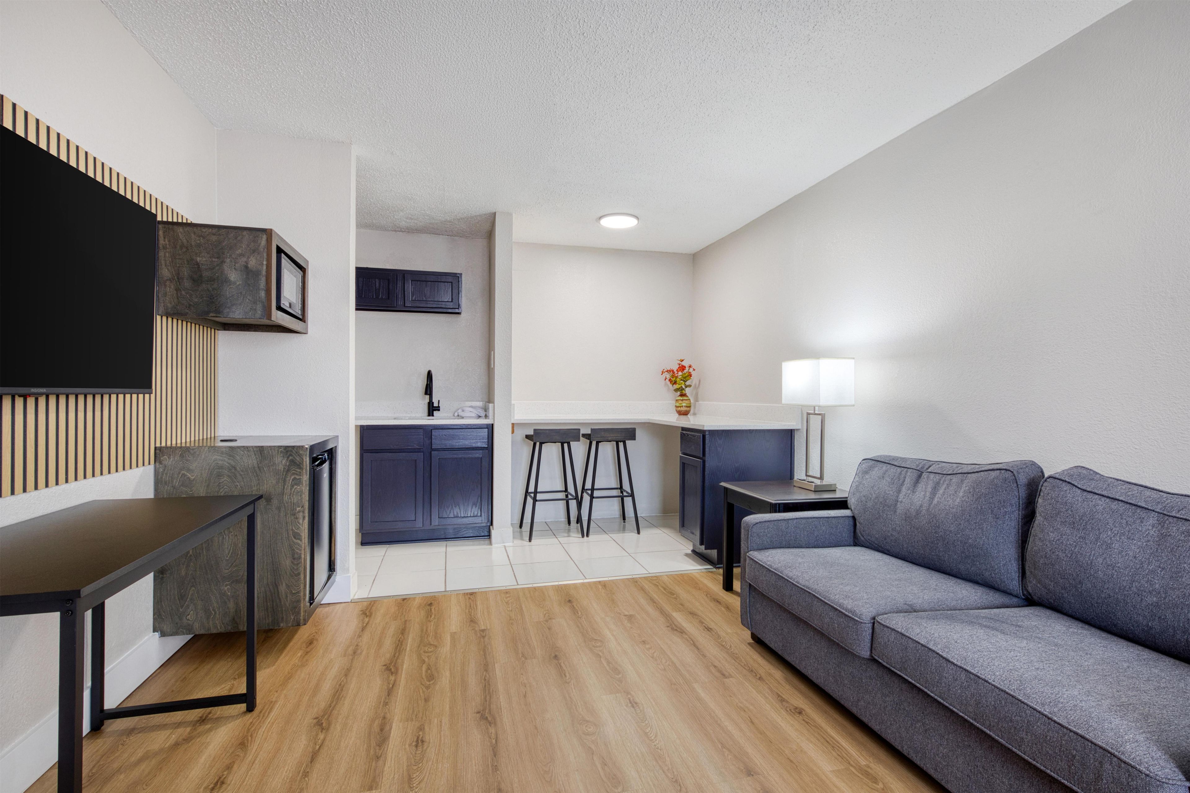 A modern living room with a grey sofa, a TV mounted on the wall, and a kitchen area with a bar stool and a vase of flowers.
