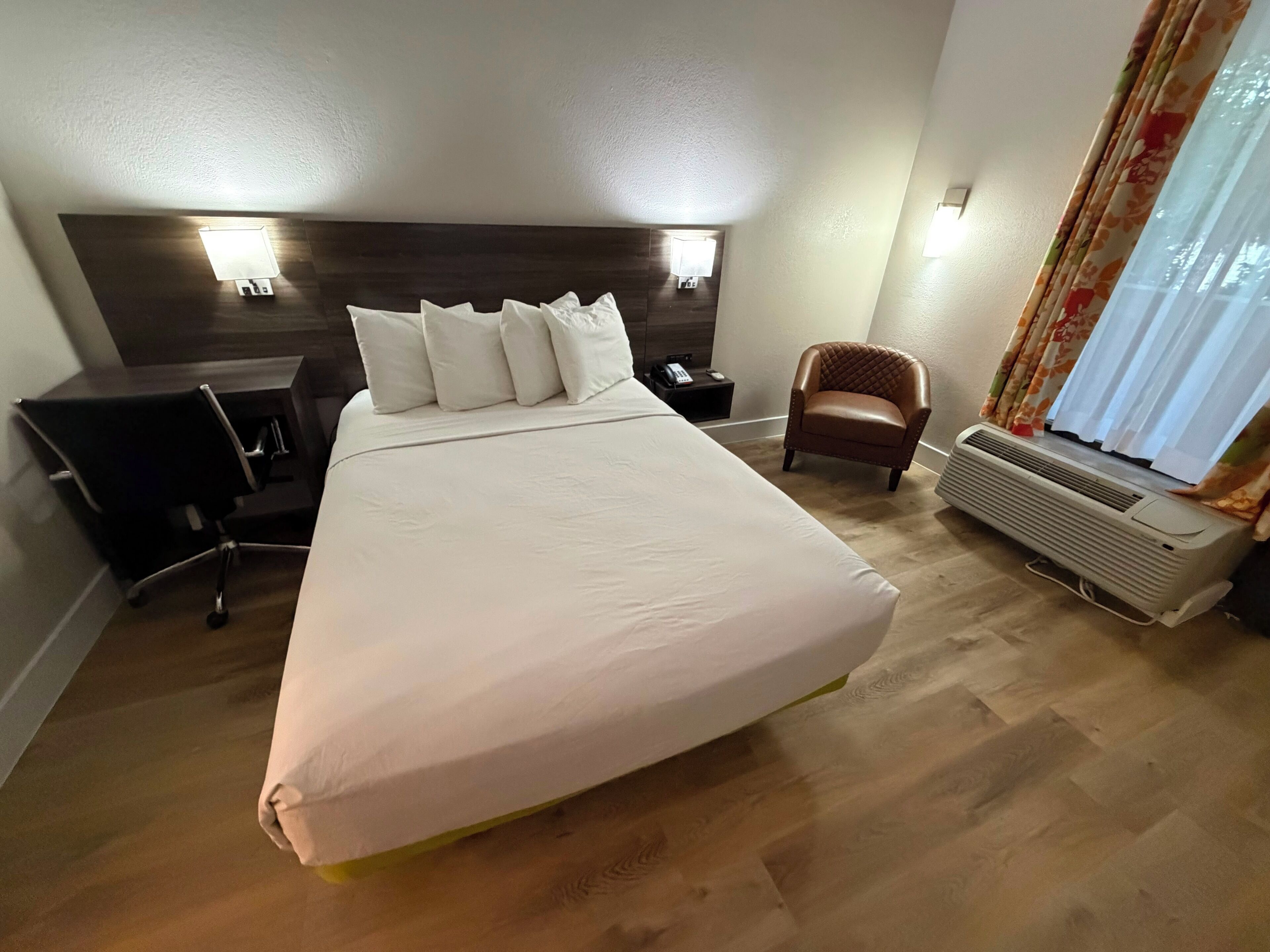 Una habitación de hotel con una cama grande, una mesita de noche con un teléfono, una silla, un escritorio y una ventana con cortinas.