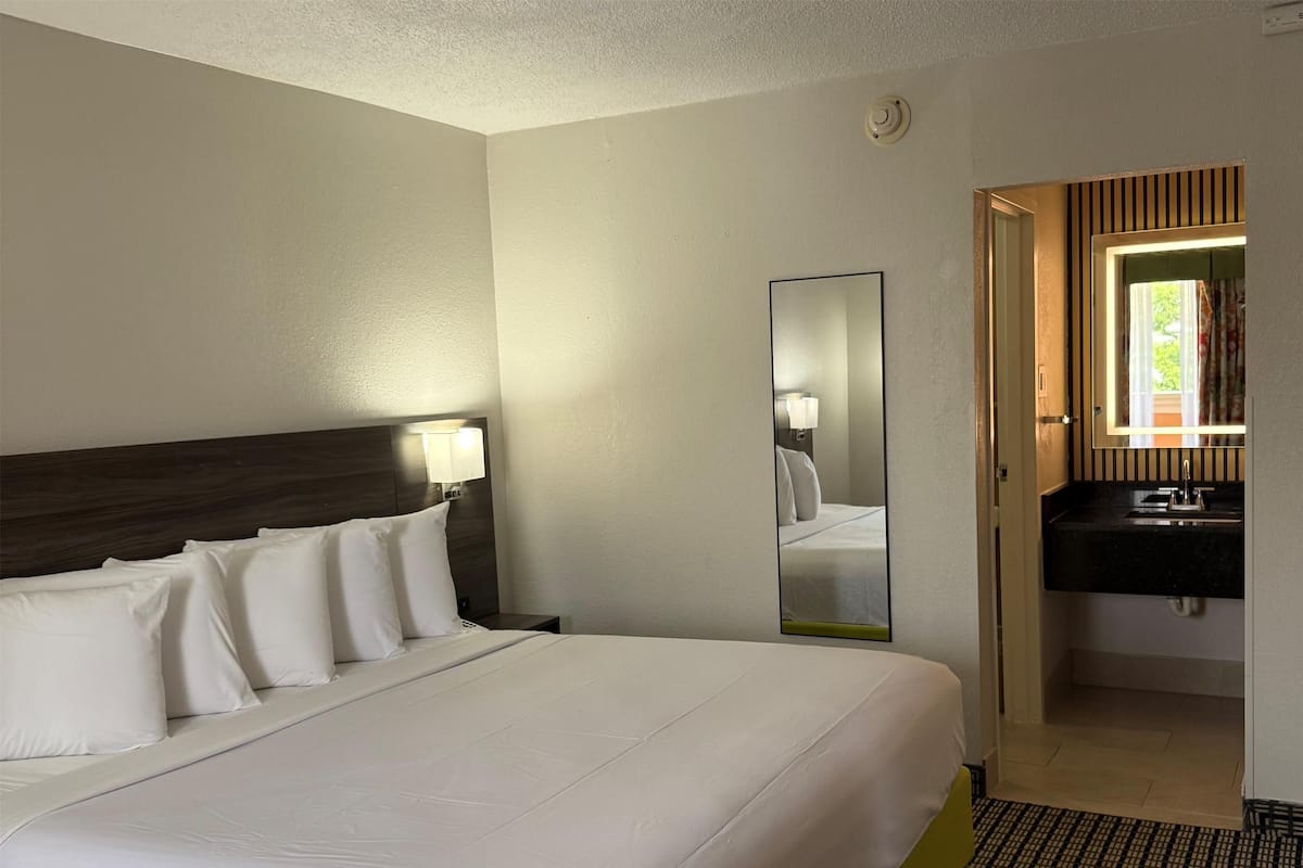 A hotel room with a large bed, a bedside table with a lamp, a mirror, and a bathroom with a sink and mirror.
