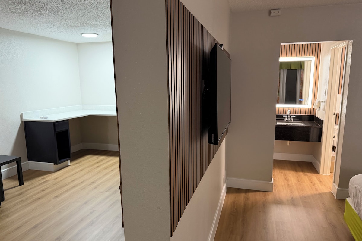 Ett modernt hotellrum med ett skrivbord placerat i hörnet, en platt-TV som är monterad på väggen, ett badrum med handfat och spegel samt trägolv.