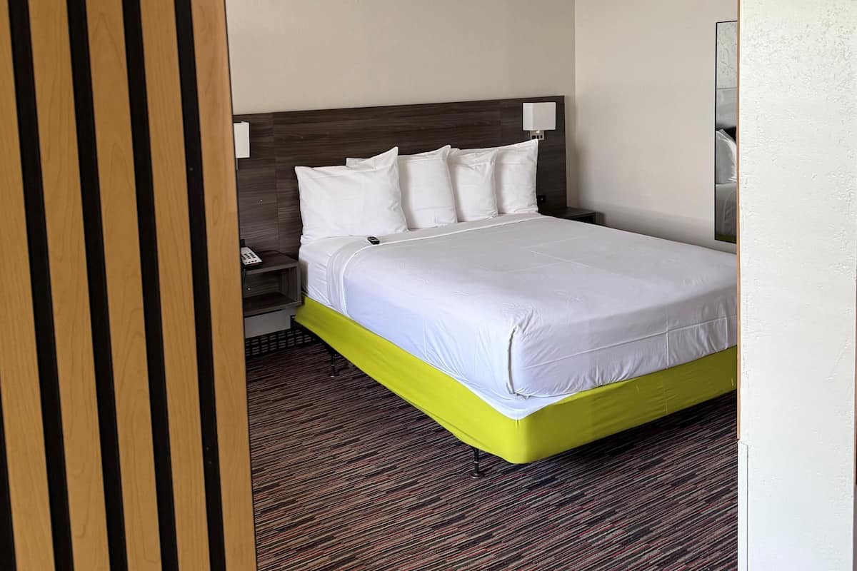 A hotel room with a bed, a nightstand, a lamp, and a wooden panel on the left.