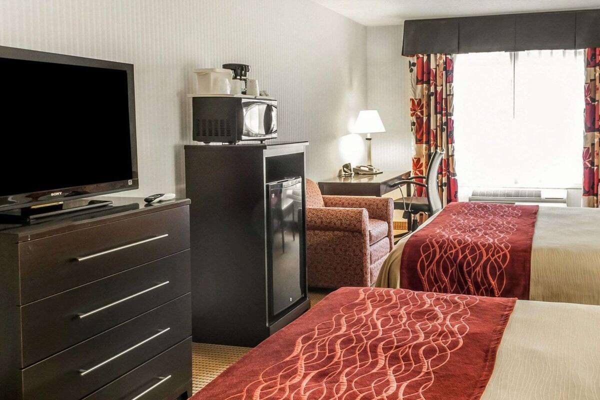 Une chambre d’hôtel équipée d’un téléviseur à écran plat, d’un réfrigérateur, d’un four à micro-ondes, d’un bureau avec une lampe, d’une chaise à motifs et de deux lits avec une literie rouge et beige.