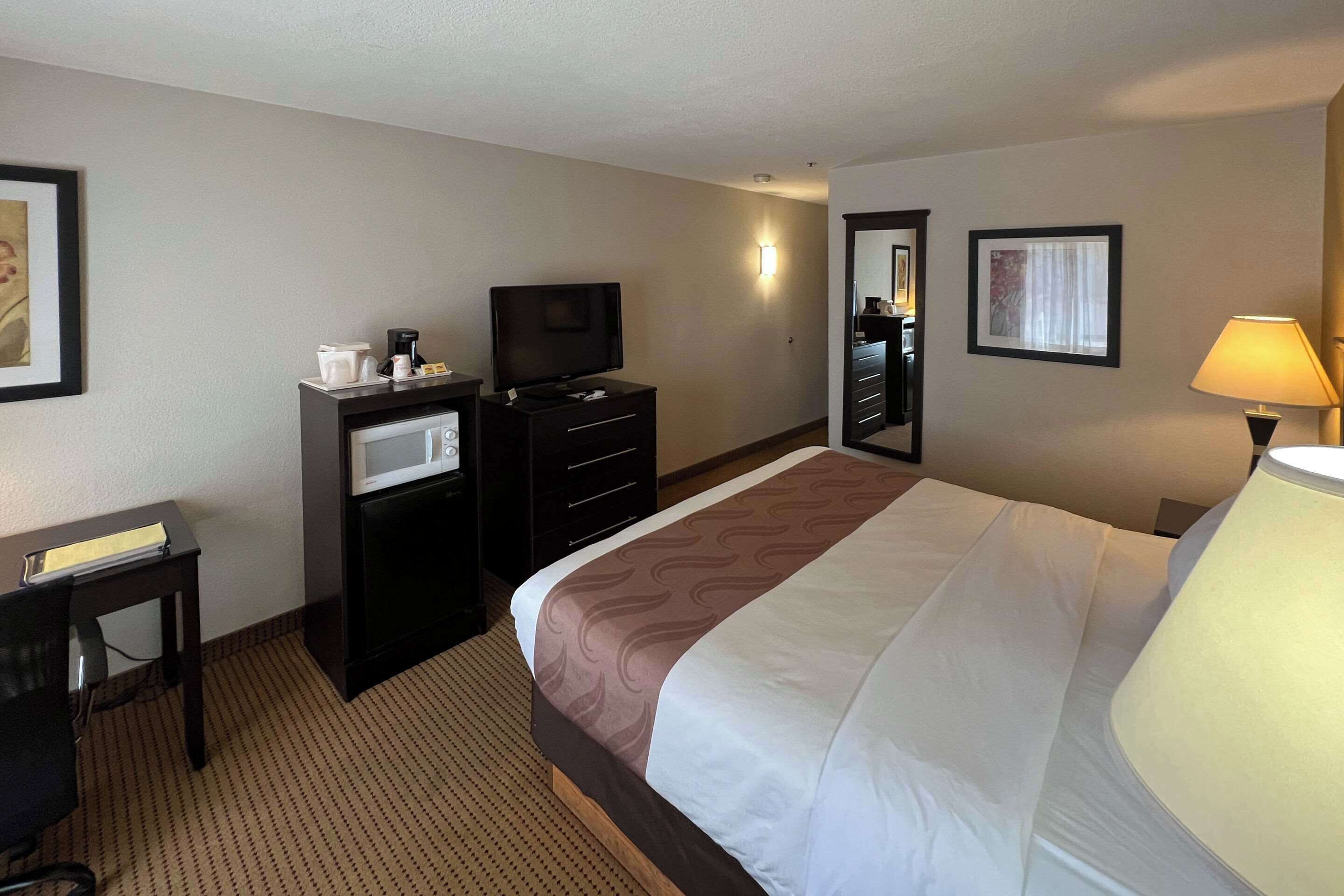 A hotel room with a bed, a TV, a desk, a lamp, and a wall-mounted light.