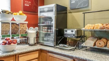 A breakfast area with a refrigerator, toaster, and various food items like fruits, cereals, and pastries.