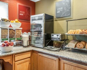 A breakfast area with a refrigerator, toaster, and various food items like fruits, cereals, and pastries.