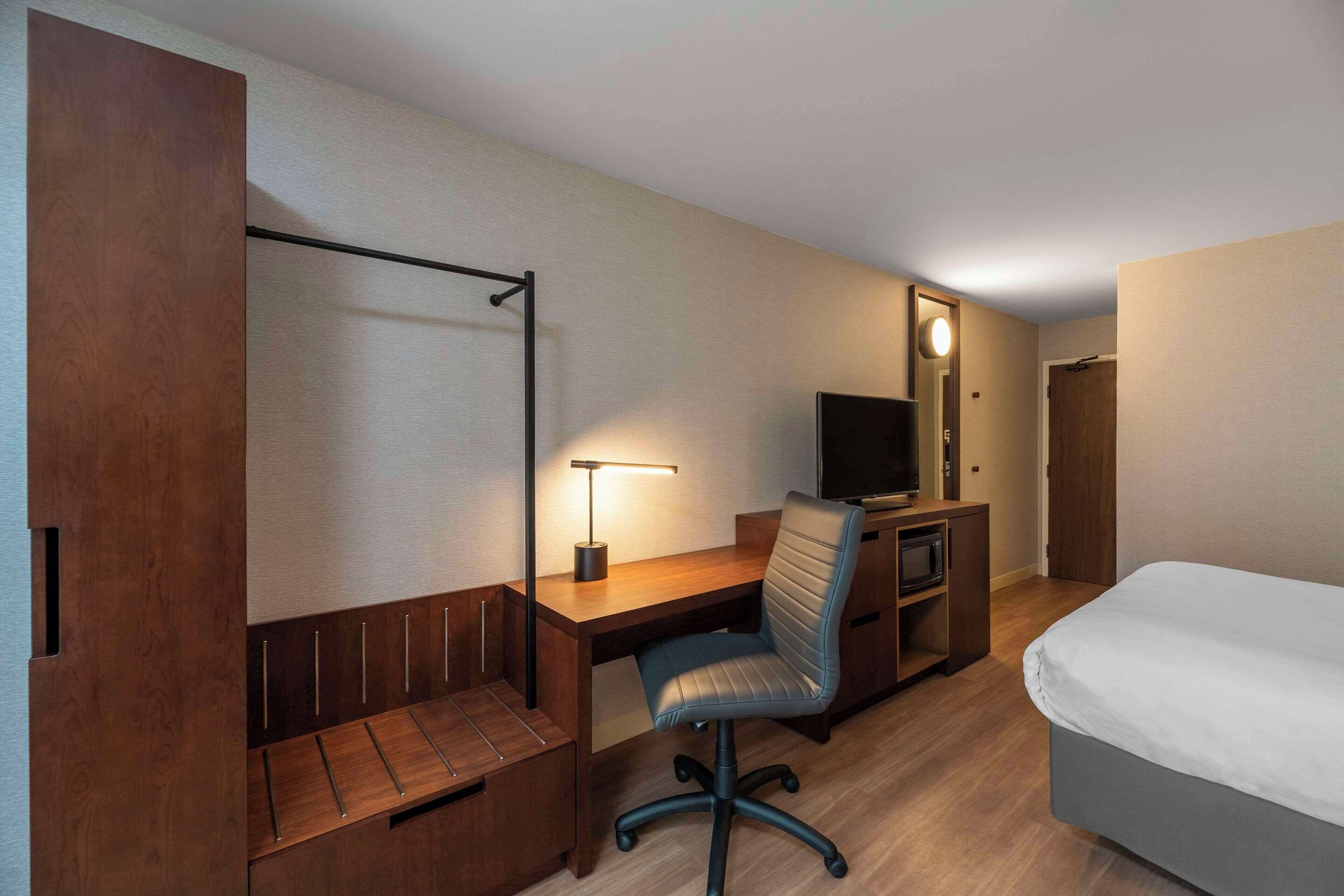 A hotel room with a wooden desk, a chair, a television, a bed, and a wardrobe.
