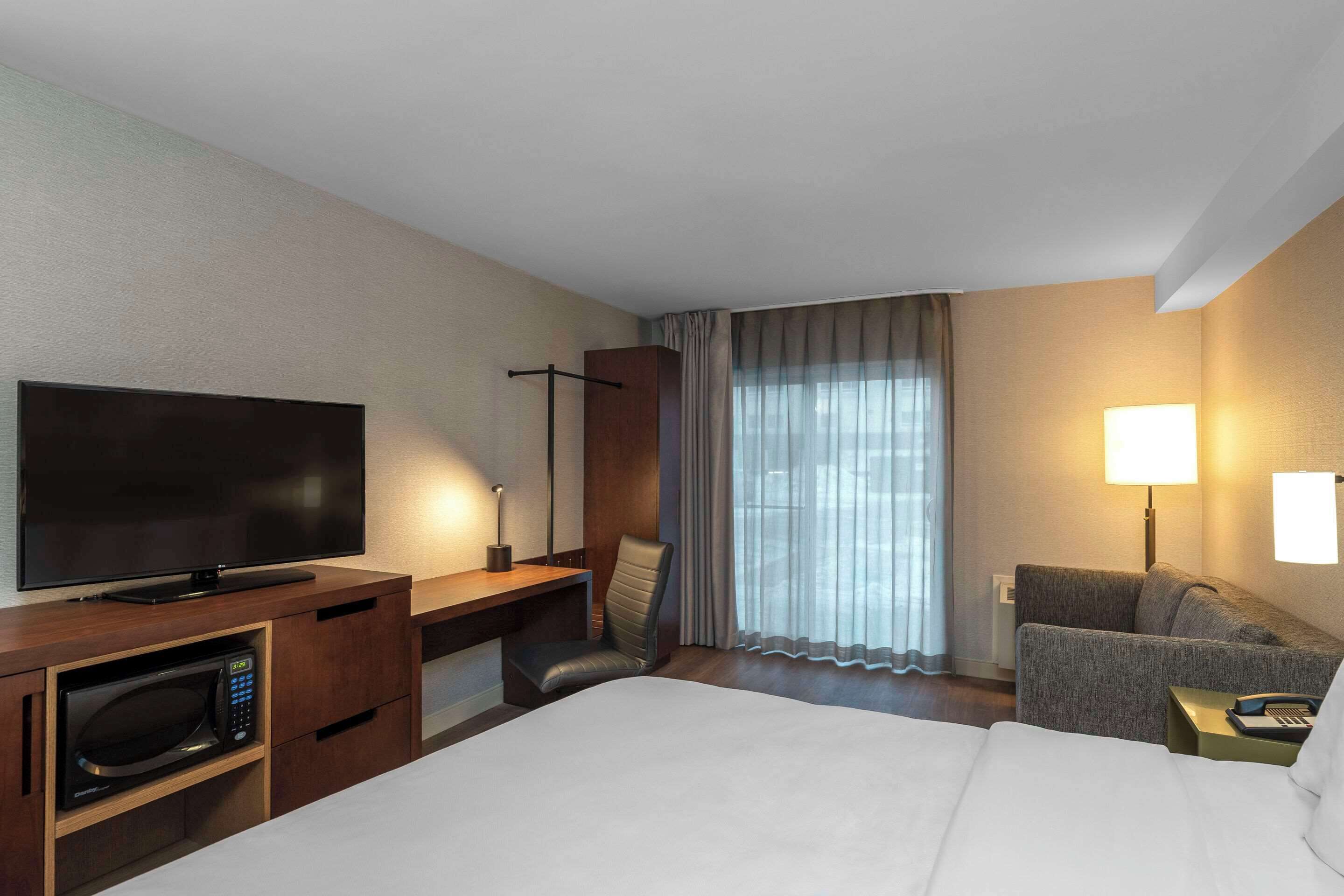 A hotel room with a bed, a TV, a desk with a chair, a microwave, a sofa, and a lamp.