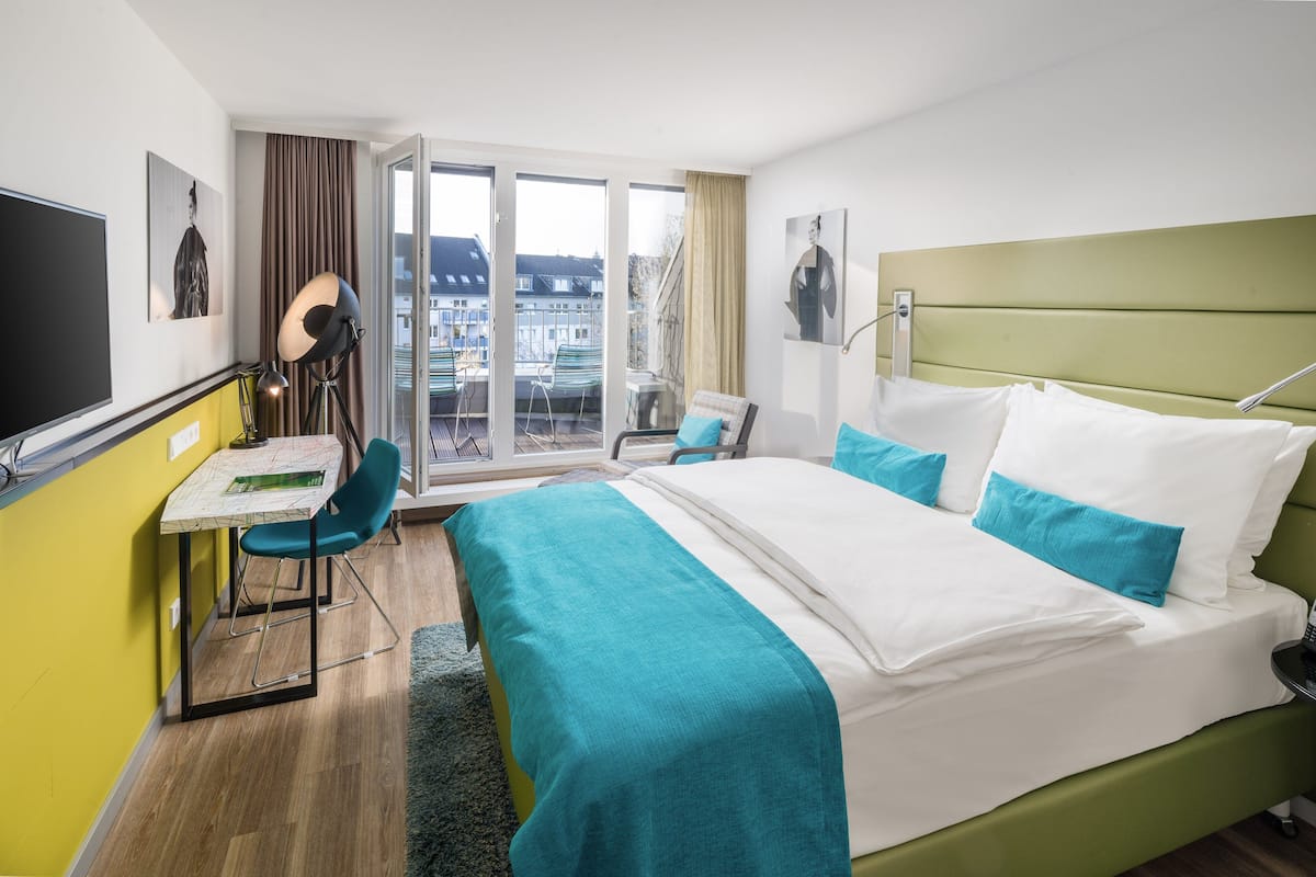 A hotel room with a bed, a desk, a chair, a TV, and a balcony with a view of buildings.