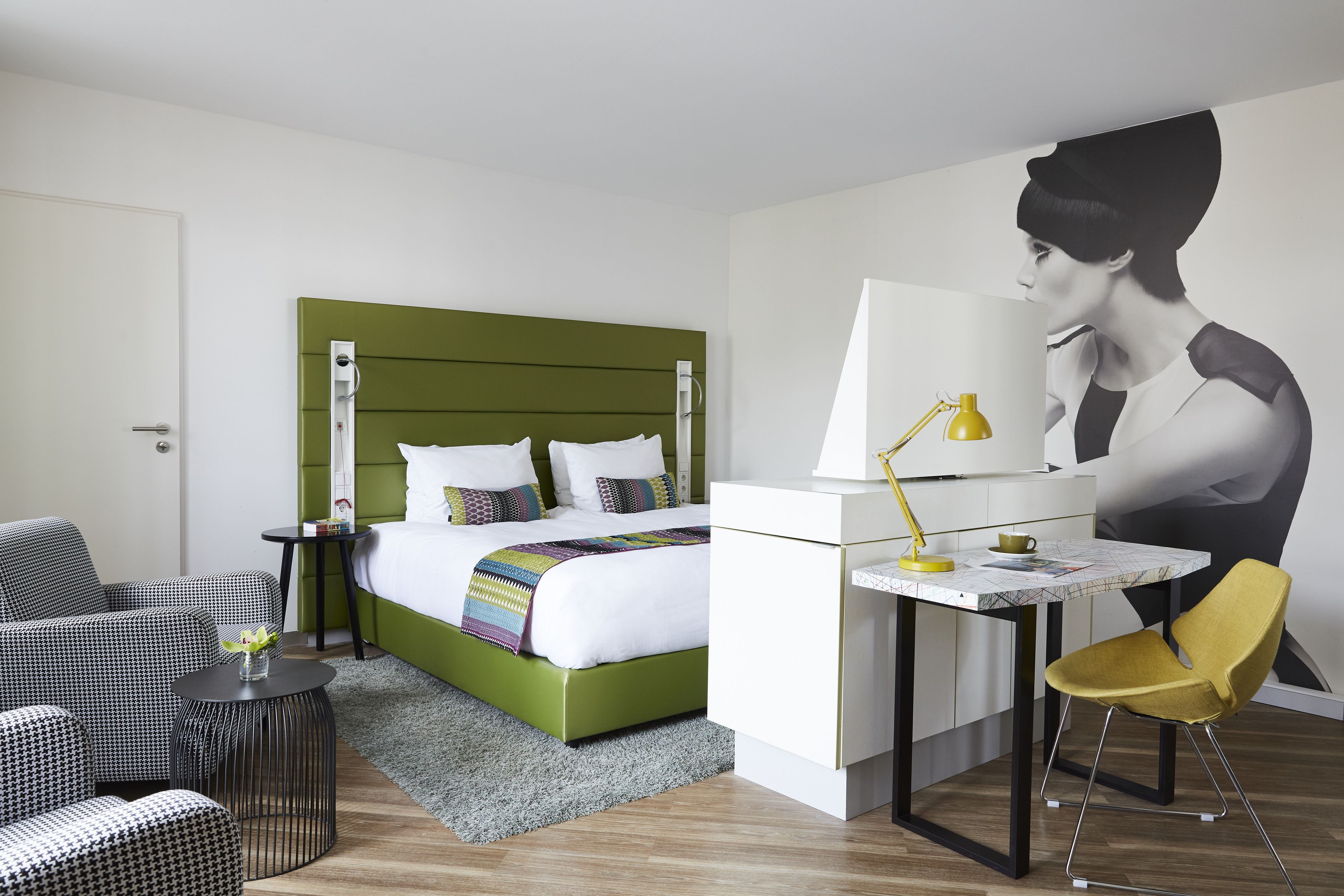 Ein Hotelzimmer mit einem grünen Bettgestell, einem Schreibtisch mit einer gelben Lampe, einem gelben Sessel und einem großen Wandbild.