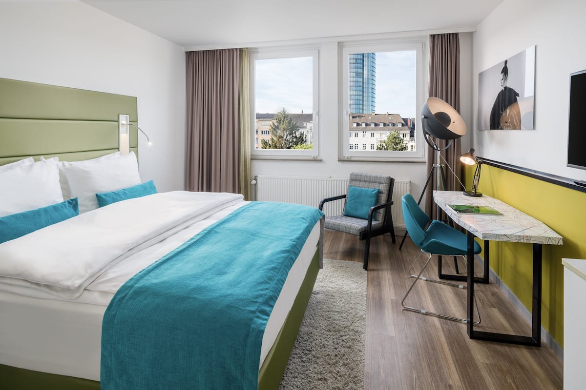 A hotel room with a bed, a desk, a chair, and a view of buildings through the window.