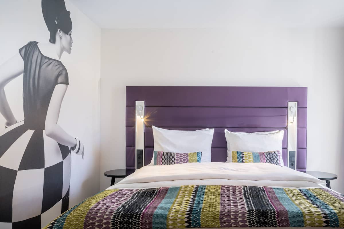 A modern hotel room with a large bed, a purple headboard, and a wall mural.