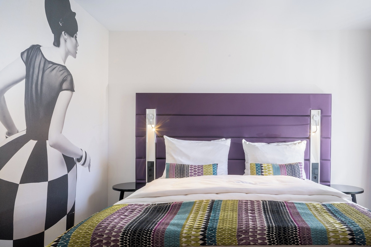 Een moderne hotelkamer met een groot bed, een paars hoofdeinde en een muurschildering.
